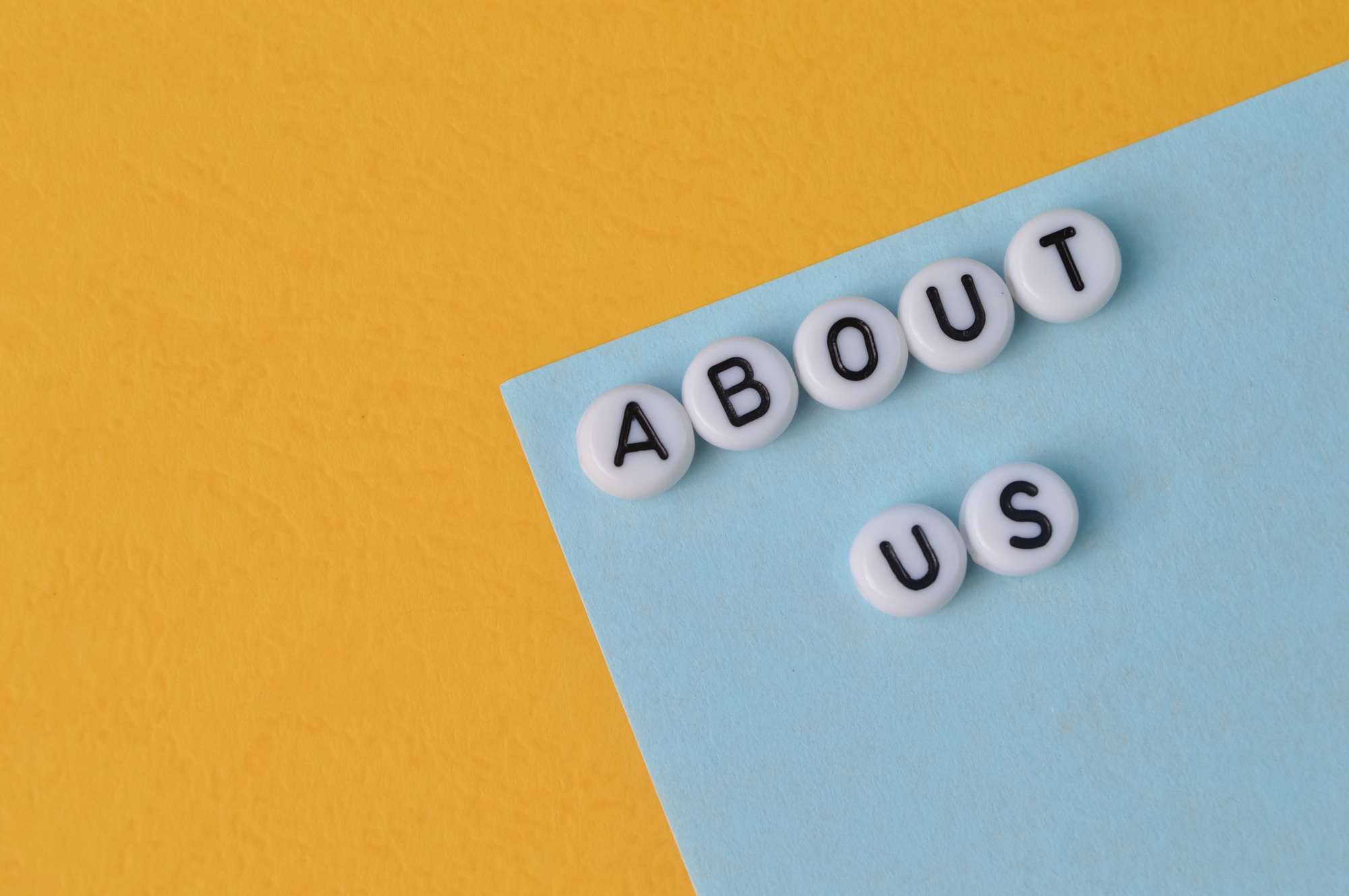 Flat lay of white letter beads spelling 'About Us' on blue and yellow background – representing healthcare practice storytelling and team introduction